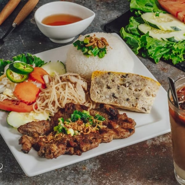 Vietnamese grilled pork, rice, egg meatloaf, and fresh vegetables on a white plate with a side of sauce.
