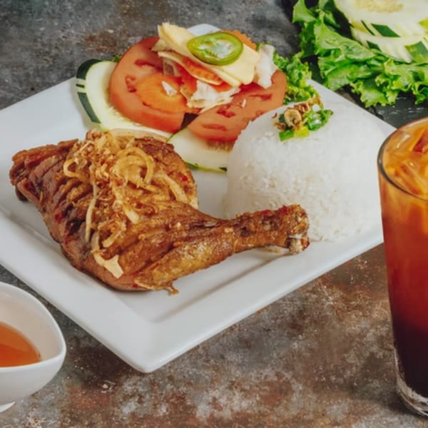 Crispy fried chicken leg with fried onions, white rice, and a side salad with tomatoes and cucumbers.