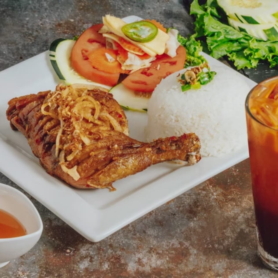Crispy fried chicken leg with fried onions, white rice, and a side salad with tomatoes and cucumbers.