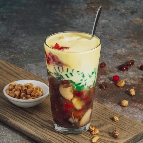 A tall glass of Vietnamese chè, a layered dessert with colorful ingredients and a spoon, next to a bowl of peanuts.