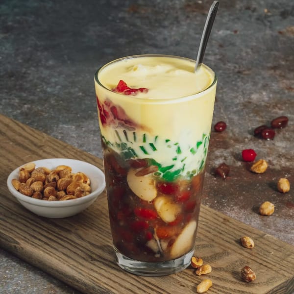 A tall glass of Vietnamese chè, a layered dessert with colorful jellies, beans, and creamy topping.