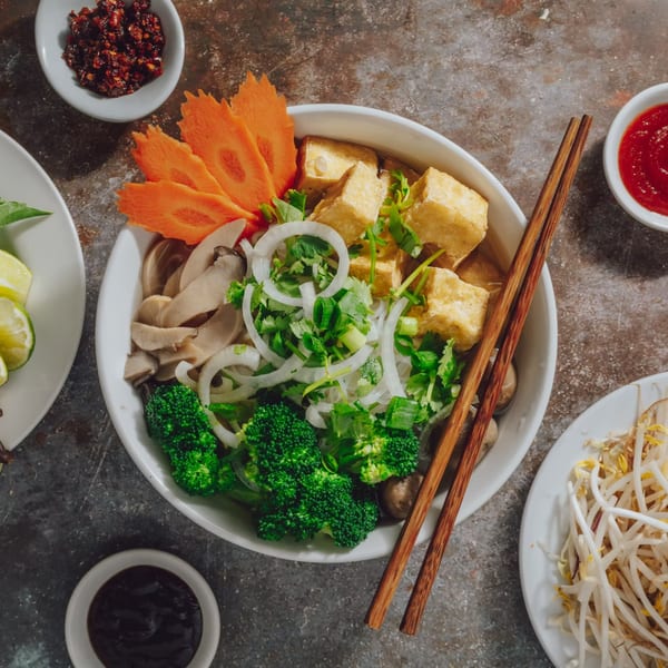 A vibrant bowl of Pho Ngon pho with tofu, broccoli, mushrooms, and carrot garnish, surrounded by sauces and bean sprouts.